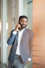 Content confident young Black businessman in jacket standing at wall and talking by mobile phone