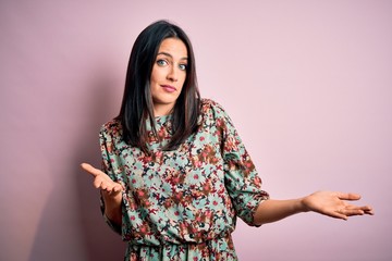 Young brunette woman with blue eyes wearing floral colorful dress over pink background clueless and...