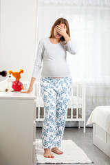 A young pregnant woman suffers from a headache and covers her eyes with her hand. Children's room in the background. The concept of the health of pregnant women, motherhood