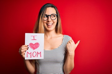 Young beautiful woman holding paper with love mom message celebrating mothers day pointing and showing with thumb up to the side with happy face smiling