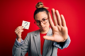 Young beautiful blonde woman with blue eyes holding reminder paper with question mark with open hand doing stop sign with serious and confident expression, defense gesture