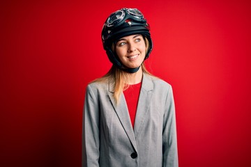 Young beautiful blonde motorcyclist woman wearing motorcycle helmet over red background looking away to side with smile on face, natural expression. Laughing confident.