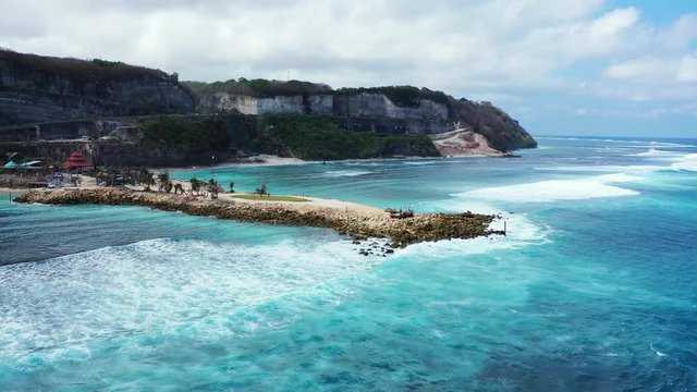 Beautiful sea scenery with white waves splashing on rocky pier seaside, slopes of green hills in Bali, Indonesia