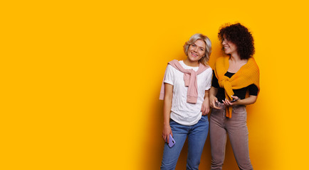 Two smiling and good looking workers are posing on a yellow background holding their phones and sharing their warm emotions