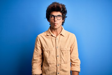 Young handsome man wearing casual shirt standing over isolated blue background making fish face with lips, crazy and comical gesture. Funny expression.
