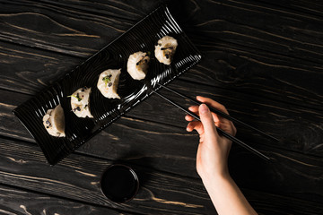 partial view of woman eating delicious Chinese boiled dumpling with chopsticks and soy sauce at black wooden table