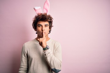 Young handsome man holding easter rabbit ears standing over isolated pink background asking to be quiet with finger on lips. Silence and secret concept.