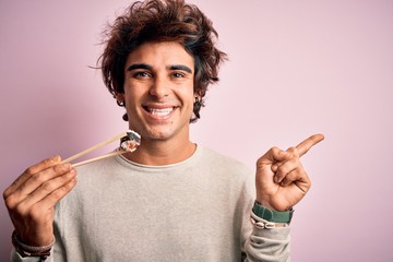 Young handsome man eating sushi using chopsticks over isolated pink background very happy pointing with hand and finger to the side
