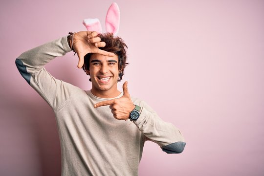 Young handsome man holding easter rabbit ears standing over isolated pink background smiling making frame with hands and fingers with happy face. Creativity and photography concept.