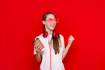 Amazed caucasian brunette teenage girl,holds coffe and wearing red earphones over red isolated...
