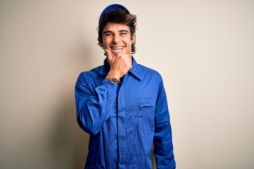 Young mechanic man wearing blue cap and uniform standing over isolated white background looking confident at the camera smiling with crossed arms and hand raised on chin. Thinking positive.