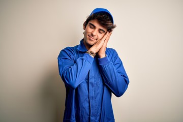Young mechanic man wearing blue cap and uniform standing over isolated white background sleeping tired dreaming and posing with hands together while smiling with closed eyes.