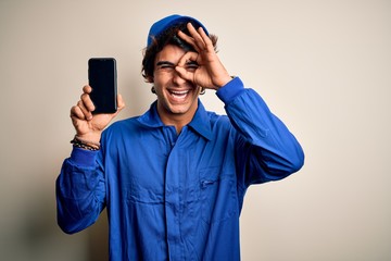 Young mechanic man wearing uniform holding smartphone over isolated white background with happy face smiling doing ok sign with hand on eye looking through fingers