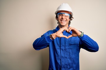 Young constructor man wearing uniform and security helmet over isolated white background smiling in love doing heart symbol shape with hands. Romantic concept.