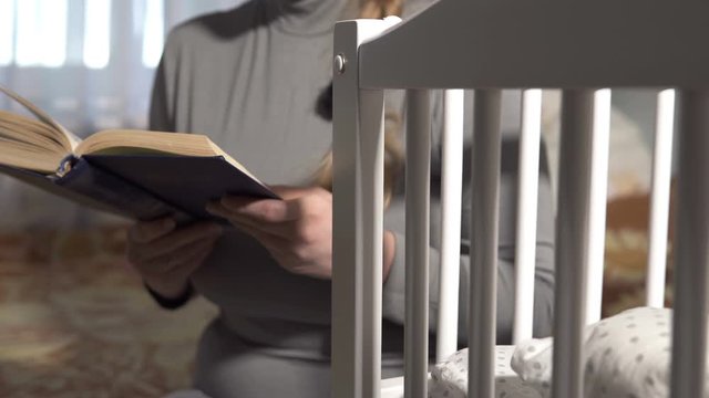 Mother is reading a book next to a sleeping baby in a crib. The concept of motherhood.