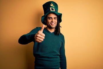 Young african american man wearing green hat with clover celebrating saint patricks day doing happy thumbs up gesture with hand. Approving expression looking at the camera showing success.