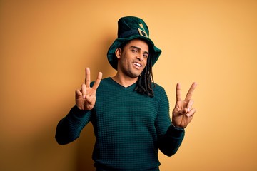 Young african american man wearing green hat with clover celebrating saint patricks day smiling looking to the camera showing fingers doing victory sign. Number two.
