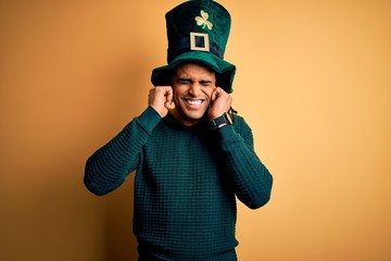 Young african american man wearing green hat with clover celebrating saint patricks day covering ears with fingers with annoyed expression for the noise of loud music. Deaf concept.
