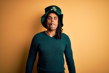 Young african american man wearing green hat with clover celebrating saint patricks day making fish face with lips, crazy and comical gesture. Funny expression.