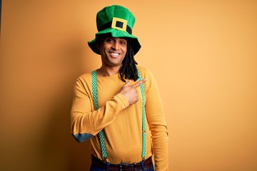 Young african american man wearing green hat celebrating saint patricks day cheerful with a smile of face pointing with hand and finger up to the side with happy and natural expression on face
