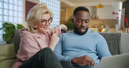 Cheerful mixed ethnicity young couple using notebook for e-commerce in living room, pensive African man and Caucasian woman doing online shopping together watching video movie resting on couch at home - Powered by Adobe