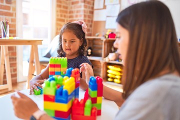Naklejka premium Beautiful teacher and toddler girl playing with construction blocks bulding tower at kindergarten