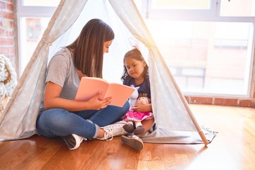 Beautiful teacher reading book to toddler girl sitting on the floor inside tipi at kindergarten