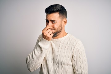 Fototapeta premium Young handsome man wearing casual sweater standing over isolated white background smelling something stinky and disgusting, intolerable smell, holding breath with fingers on nose. Bad smell