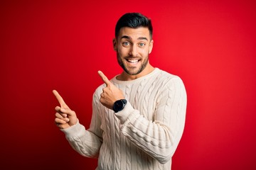Young handsome man wearing casual white sweater standing over isolated red background smiling and looking at the camera pointing with two hands and fingers to the side.