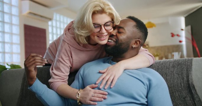 Cute Caucasian woman coming from back to hug and kiss boyfriends sitting on couch at home, pensive man smiling looking at beautiful blonde girlfriend holding mobile phone resting in living room