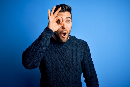 Young handsome man wearing casual sweater standing over isolated blue background doing ok gesture shocked with surprised face, eye looking through fingers. Unbelieving expression.