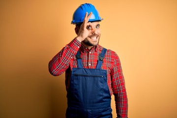 Young builder man wearing construction uniform and safety helmet over yellow background doing ok gesture with hand smiling, eye looking through fingers with happy face.
