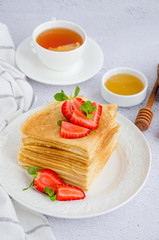 Crepes, traditional Russian thin pancakes on a white plate with fresh strawberries and honey on a light background. Russian maslenitsa. Vertical orientation. Close up.