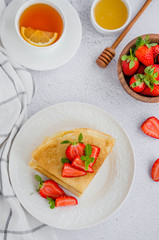 Crepes, traditional Russian thin pancakes on a white plate with fresh strawberries and honey on a light background. Russian maslenitsa. Vertical orientation. Top view