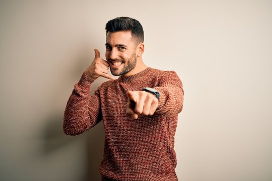 Young Handsome Man Wearing Casual Sweater Standing Over Isolated White Background Smiling Doing Talking On The Telephone Gesture And Pointing To You. Call Me.