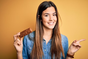 Young beautiful girl holding healthy protein bar standing over isolated yellow background very...