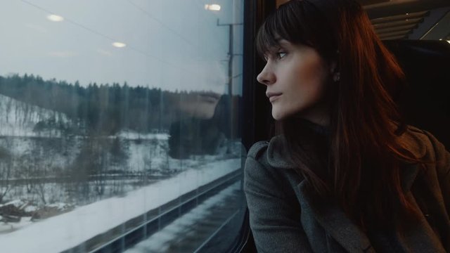 Portrait of thoughtful young attractive European woman looking out of moving winter train on window seat slow motion.