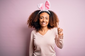 Obraz na płótnie Canvas Young african american woman with afro hair wearing bunny ears over pink background showing and pointing up with finger number one while smiling confident and happy.