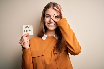 Young beautiful blonde woman holding pay taxes to goverment reminder over yellow background with happy face smiling doing ok sign with hand on eye looking through fingers