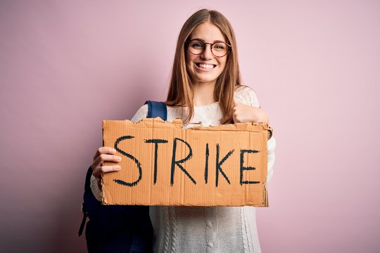 Young beautiful redhead activist woman holding banner with strike message with surprise face pointing finger to himself