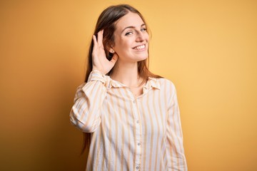 Young beautiful redhead woman wearing casual striped shirt over isolated yellow background smiling with hand over ear listening an hearing to rumor or gossip. Deafness concept.