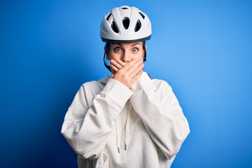 Young beautiful redhead cyclist woman wearing bike helmet over isolated blue background shocked covering mouth with hands for mistake. Secret concept.