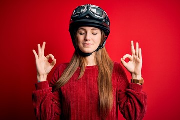 Young beautiful redhead motocyclist woman wearing moto helmet over red background relax and smiling with eyes closed doing meditation gesture with fingers. Yoga concept.
