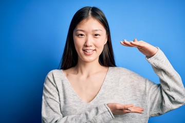 Young beautiful asian woman wearing casual sweater standing over blue isolated background gesturing with hands showing big and large size sign, measure symbol. Smiling looking at the camera. Measuring