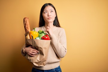Young asian woman holding paper bag of fresh healthy groceries over yellow isolated background Thinking concentrated about doubt with finger on chin and looking up wondering