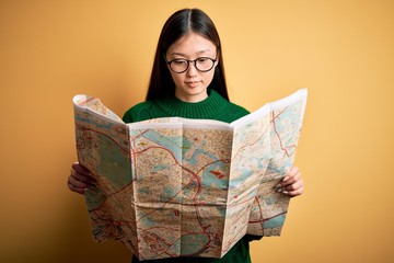 Young asian turist woman looking at city tourist map on a trip over yellow background with a confident expression on smart face thinking serious