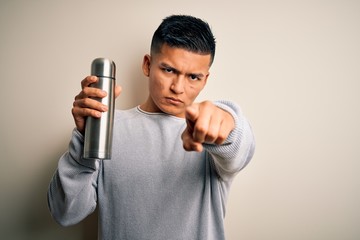 Young handsome latin man holding thermo with water over isolated white background pointing with finger to the camera and to you, hand sign, positive and confident gesture from the front