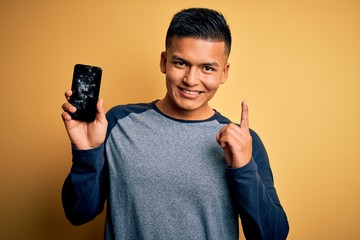 Young handsome latin man holding broken smartphone showing craked screen surprised with an idea or...