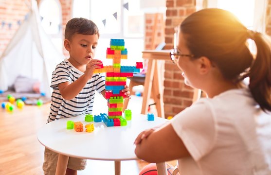 Beautiful teacher and toddler boy playing with construction blocks bulding tower at kindergarten
