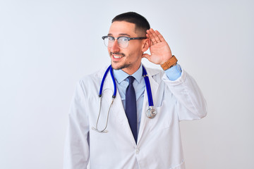 Young doctor man wearing stethoscope over isolated background smiling with hand over ear listening an hearing to rumor or gossip. Deafness concept.